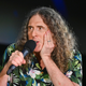An image of Weird Al Yankovic with a microphone to his month and holding his face.
